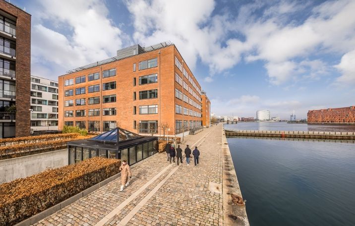 Personer går langs en havn i en by, med bygninger og kanaler i baggrunden