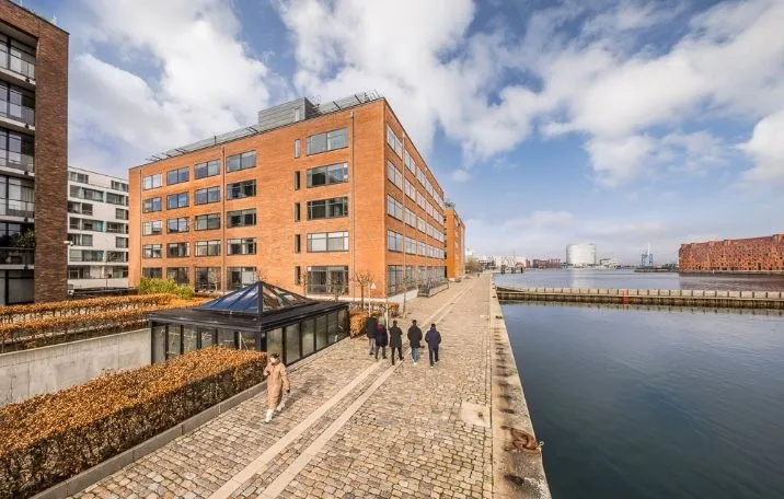 Personer går langs en havn i en by, med bygninger og kanaler i baggrunden