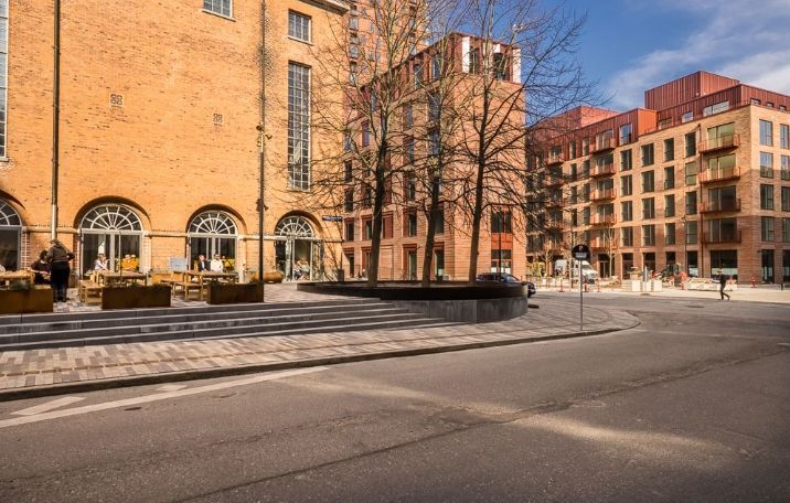 Et moderne kontorbyggeri med røde mursten og glasvinduer og et stort rekreativt område med træer og møbelsæt med mennesker