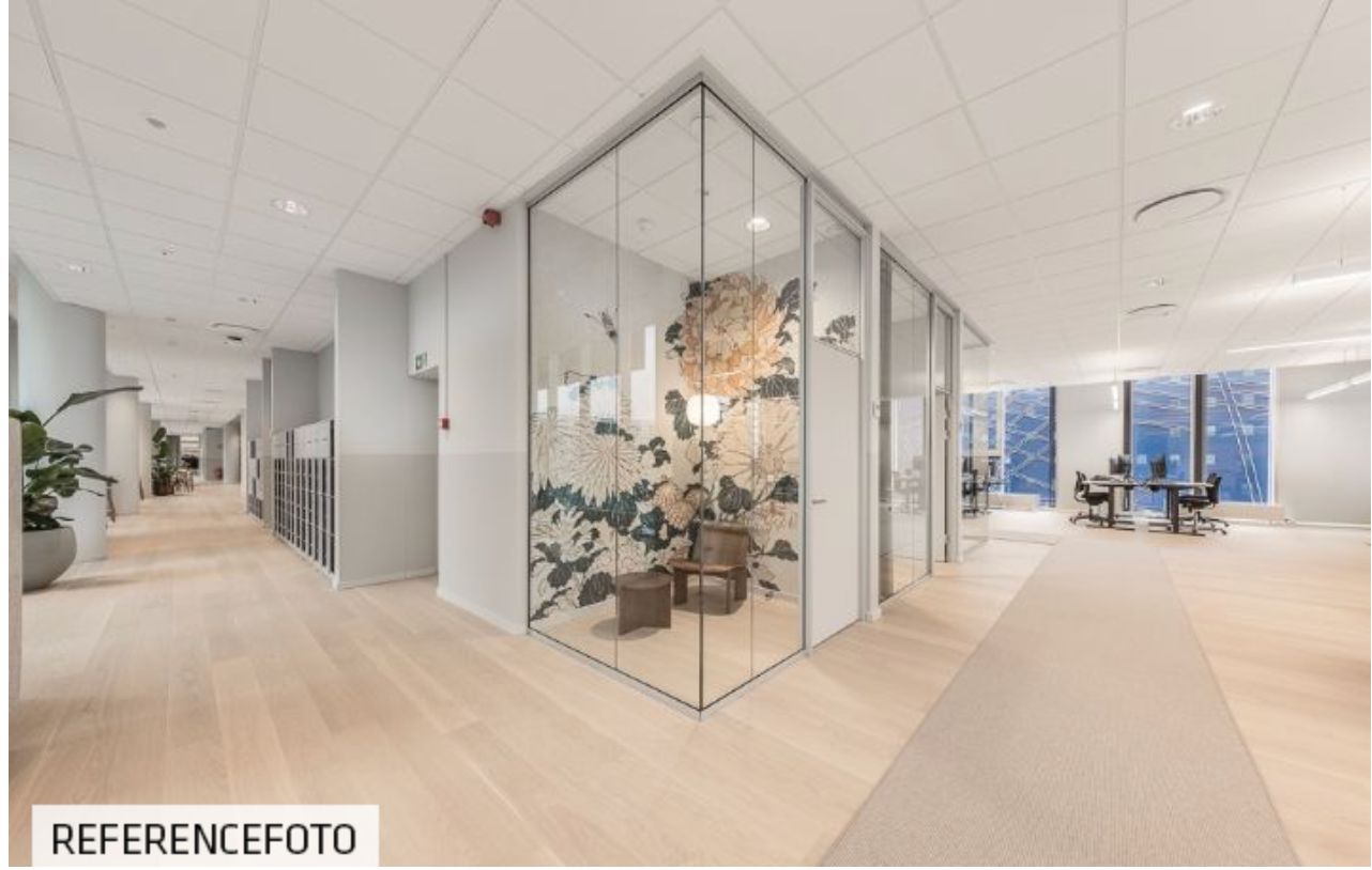 Inde i en stor kontorbygning med lyse gulve og hvide lofter er der en glasdør og et møbelstykke med en plante og lyse møbler