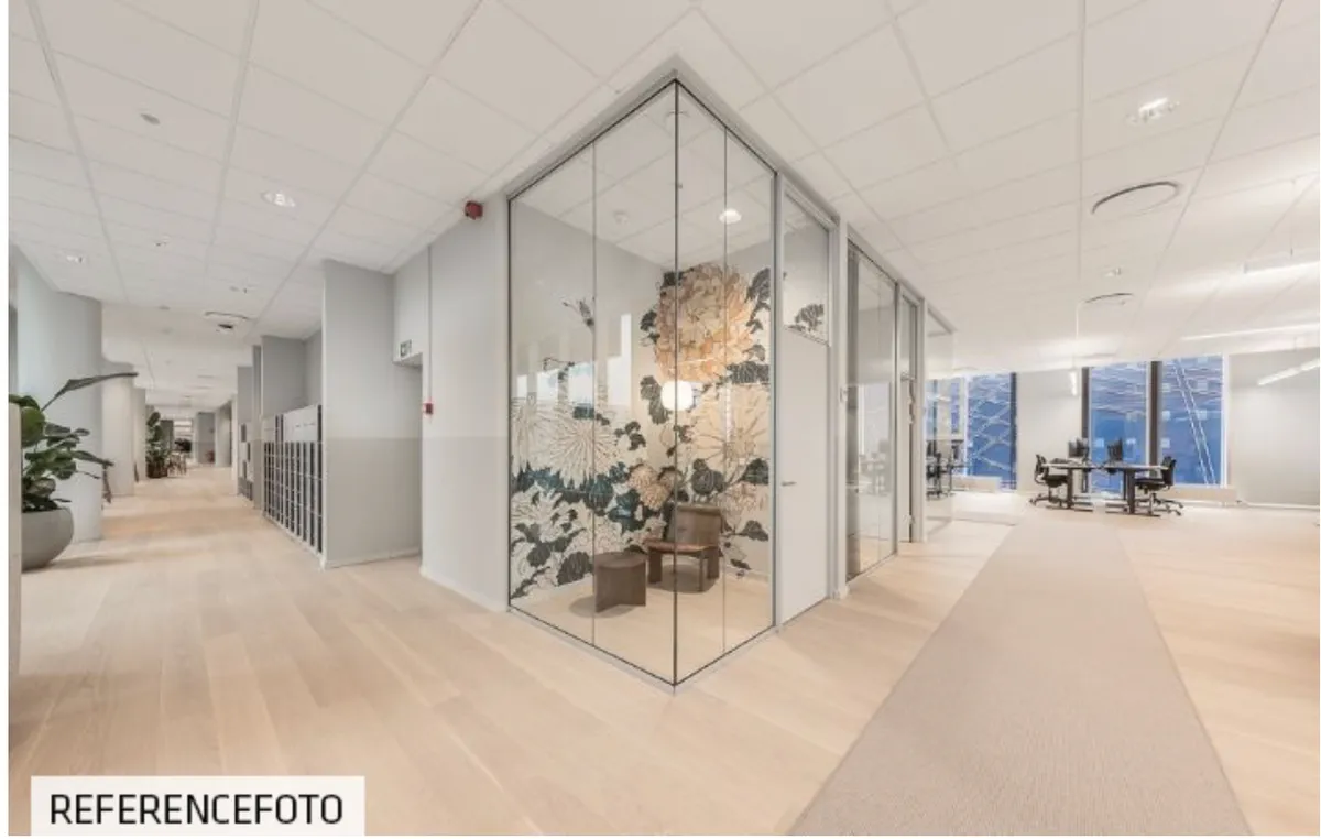 Inde i en stor kontorbygning med lyse gulve og hvide lofter er der en glasdør og et møbelstykke med en plante og lyse møbler