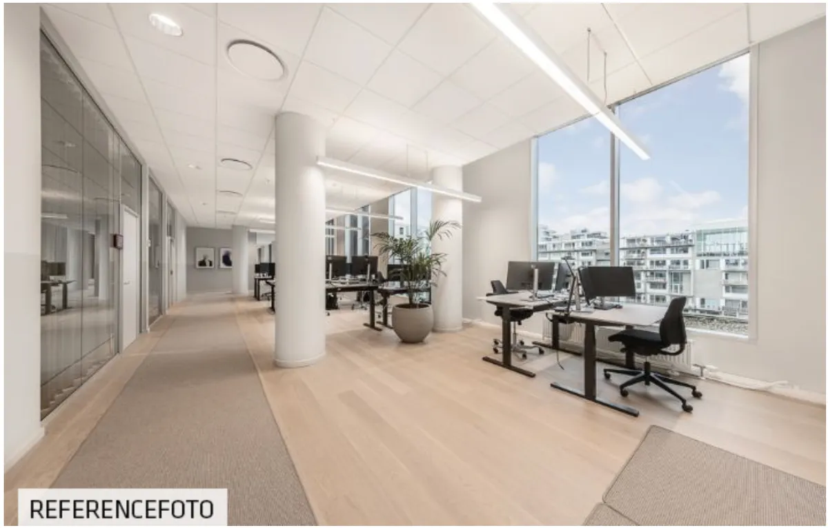 Moderne kontor med lyse lofter, gulv og glasvægge. To skriveborde med computere og en potteplante. Moderne møbler og lyse