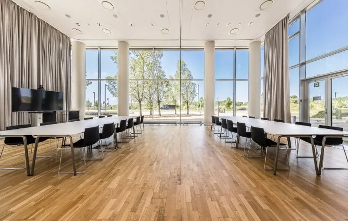 Modern konferencelokale med trægulv, store vinduer, lange borde og stole samt en stor skærm