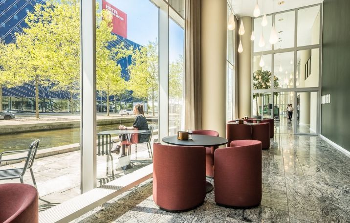 Kvinde sidder ved bord og drikker i lobby med moderne møbler og glasvægge