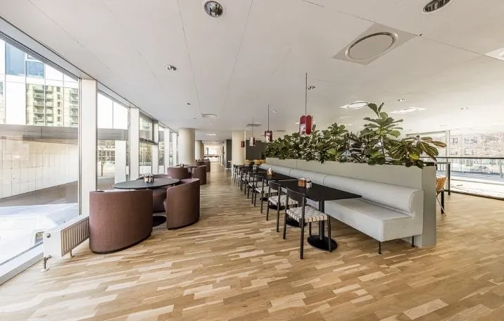 En stor og moderne kontorlounge med trægulve, runde borde og stole samt lyse glasruder og planter