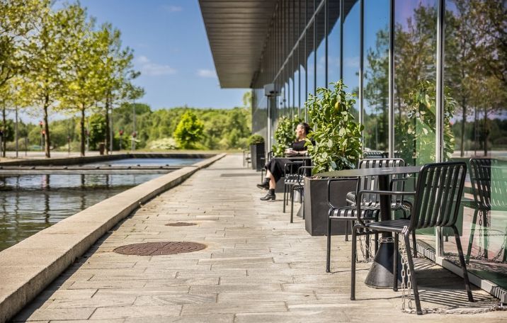 En kvinde sidder og læser på en terrasse i et kontorområde med udsigt over en sø og smukke grønne omgivelser