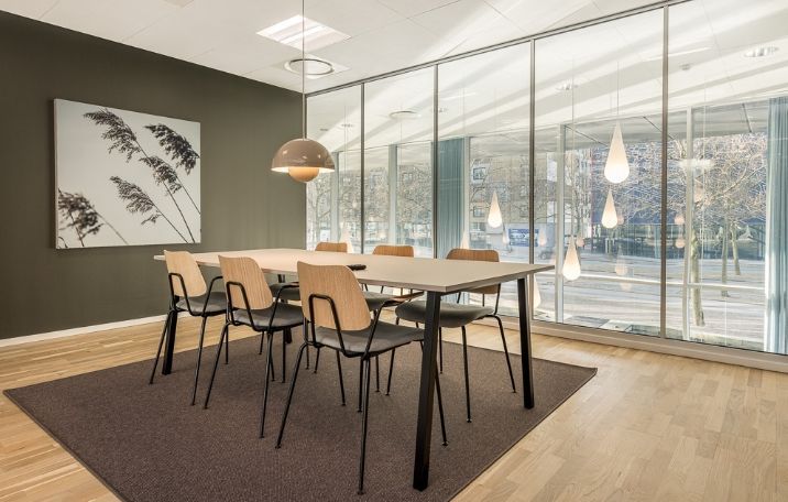 Minimalistisk kontor med lyse trægulve, moderne møbler og store glasruder mod en grøn park og en vej