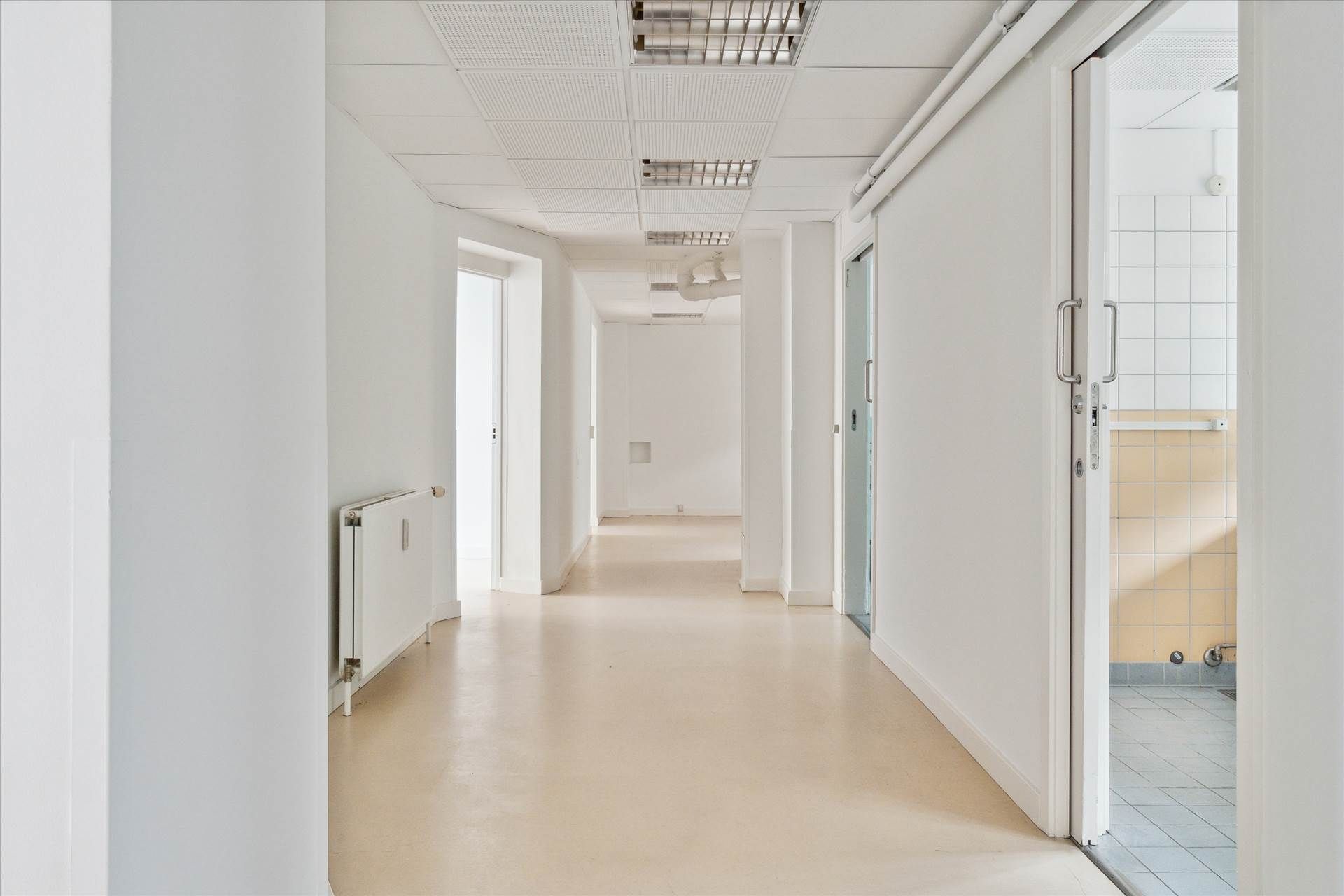 En lang og lys hall med hvide vægge og gulv samt åbne døre mod toilet og kontor