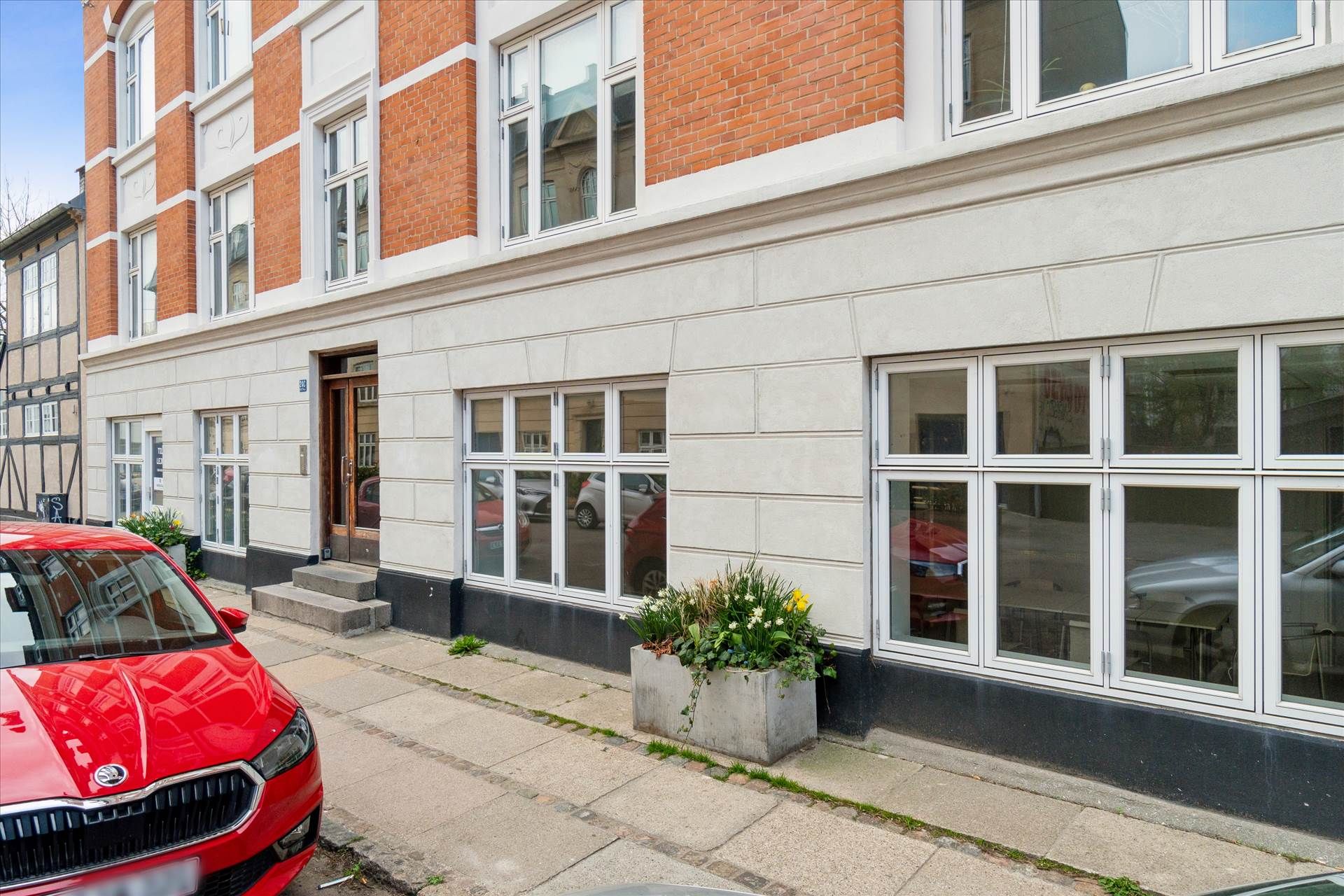 En rødt Skoda-bil parkeret foran en bygning med hvide og røde mursten og store vinduer samt en plantekasse med blomster