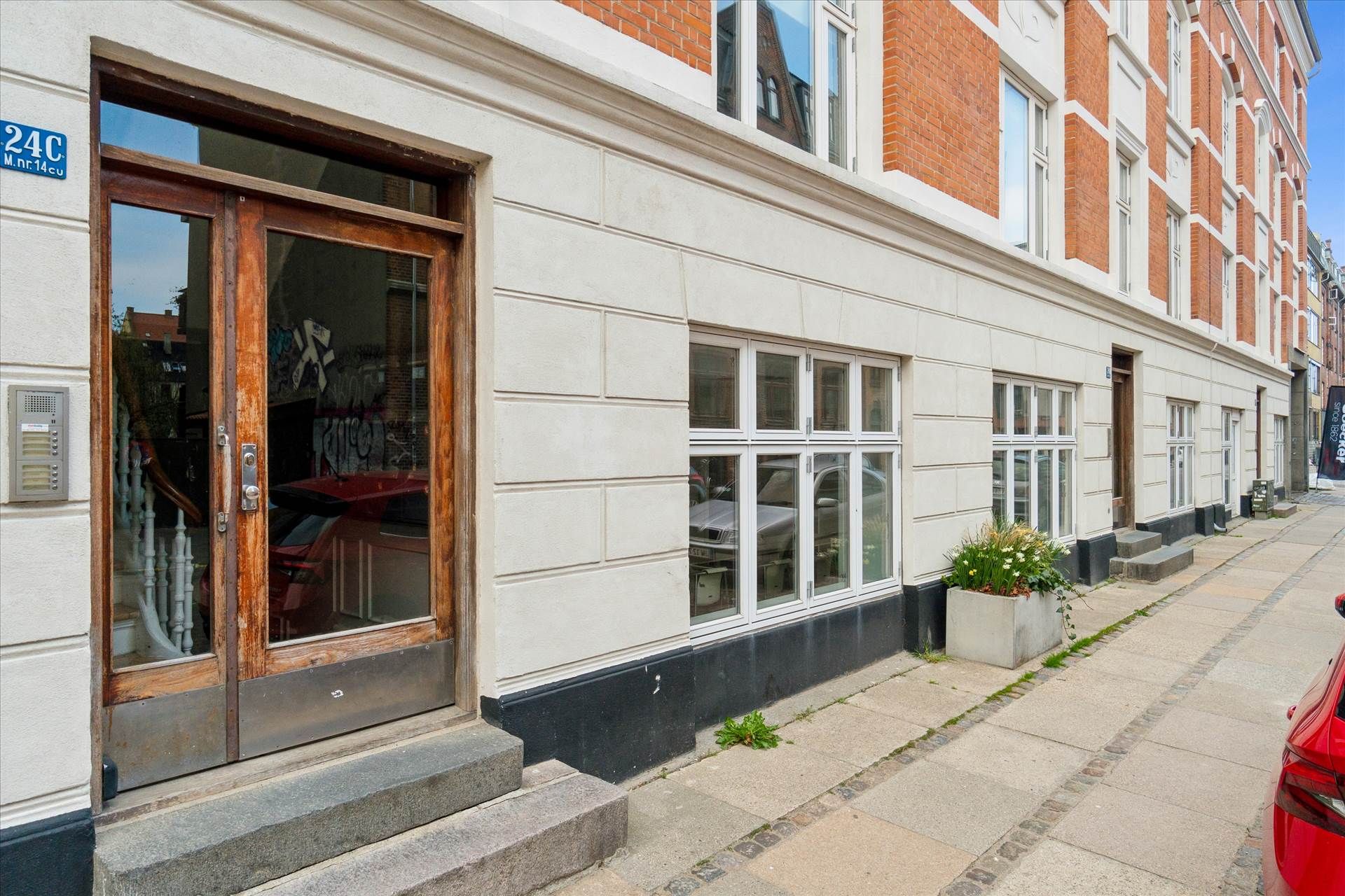 Lejlighed med glasdøre og terrassedøre. En rødfarvet bil parkeret foran døren. Et gulvplante i en potte. En hvid bygning med