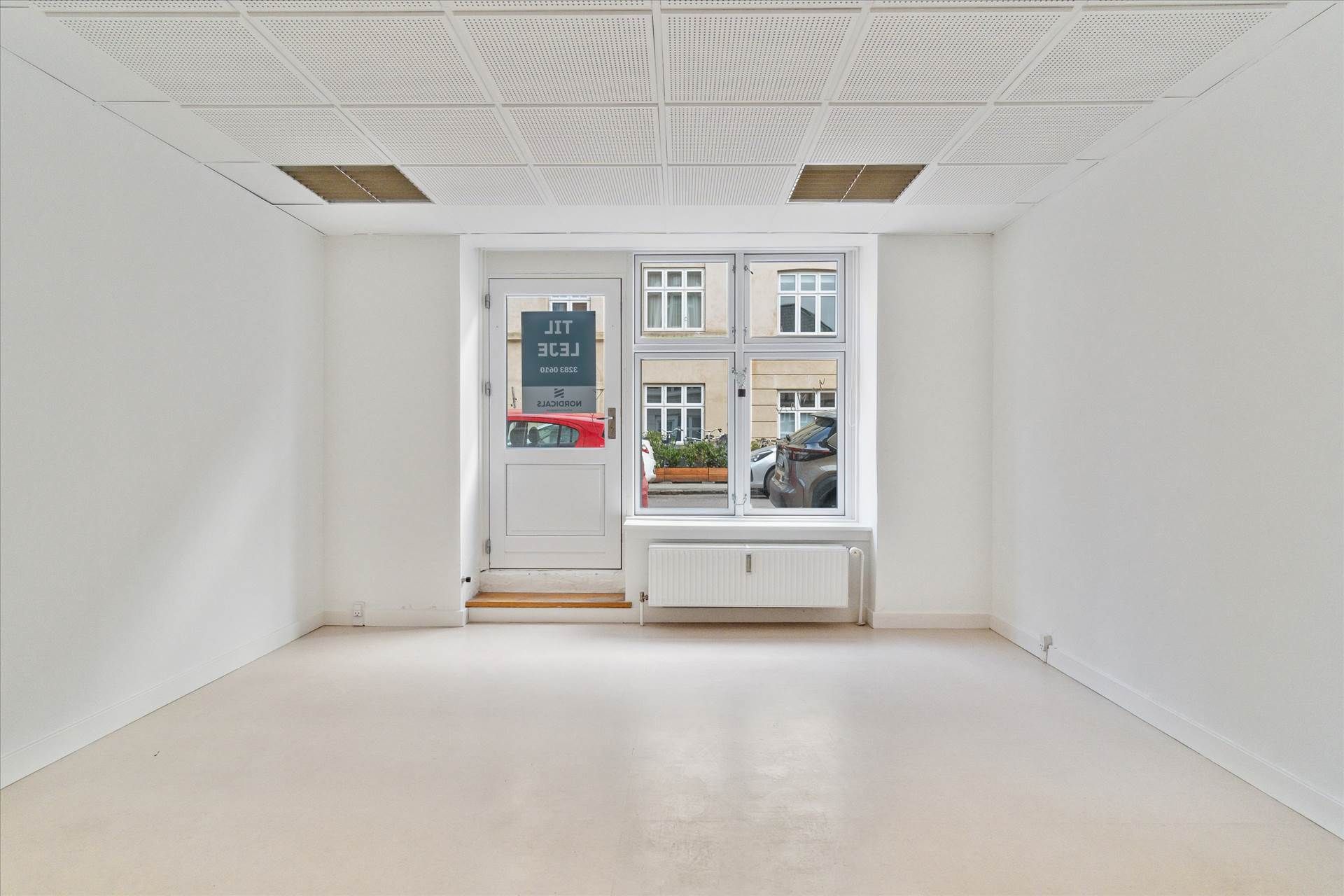 Lejlighed med hvide vægge og gulv samt glasdør og vinduer med biler og bygninger udenfor