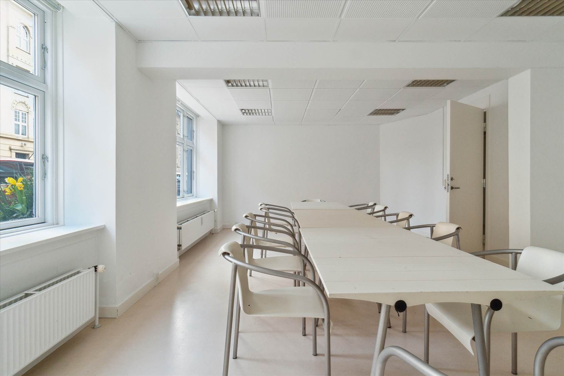 Et stort rum med hvide vægge, hvide borde og stole samt radiatorer og vinduer med udsigt til gården