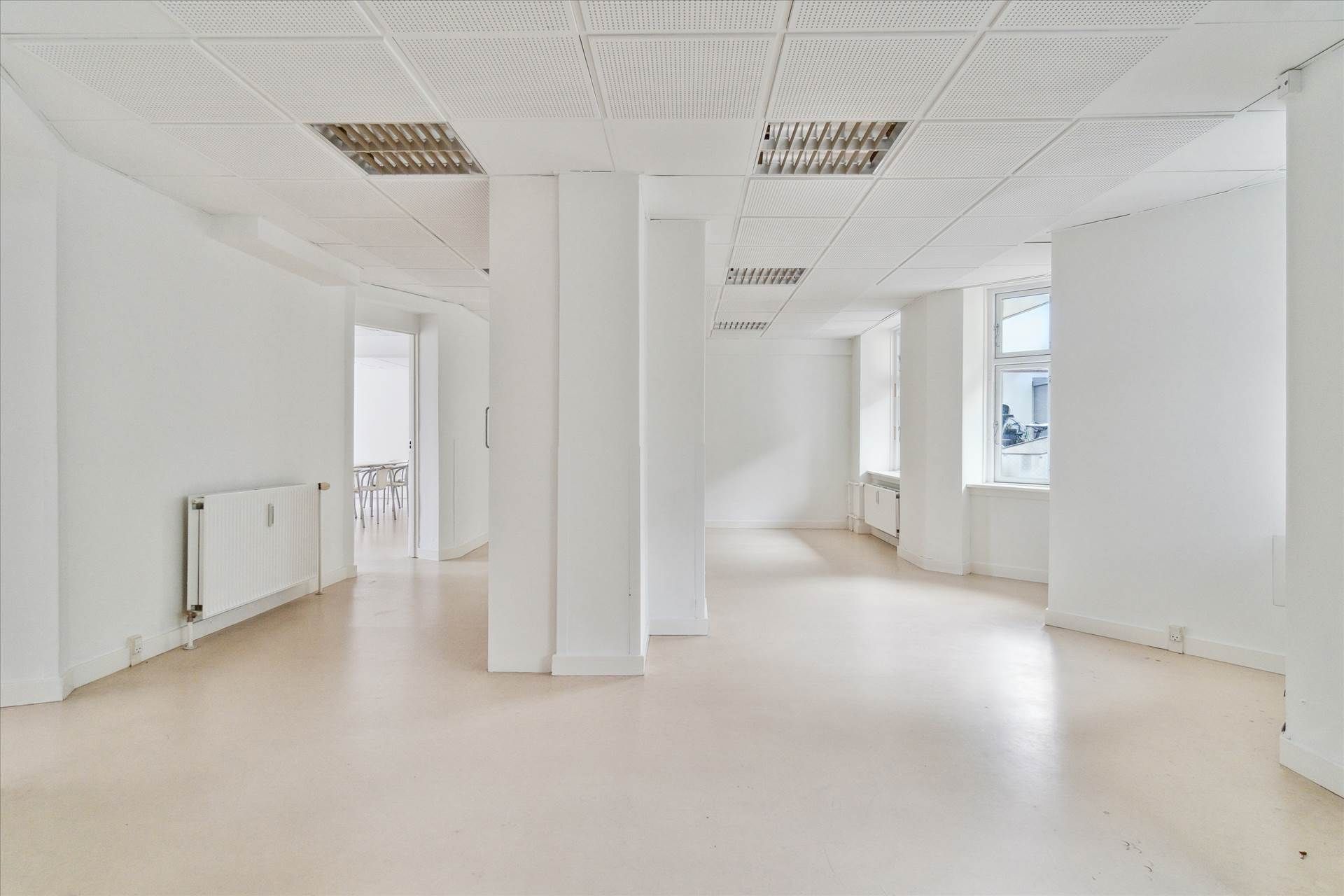 Lys og stor kælderlejlighed med højt til loftet og hvide vægge samt gulv. Rummet er møbleret med kontormøbler og radiatorer.