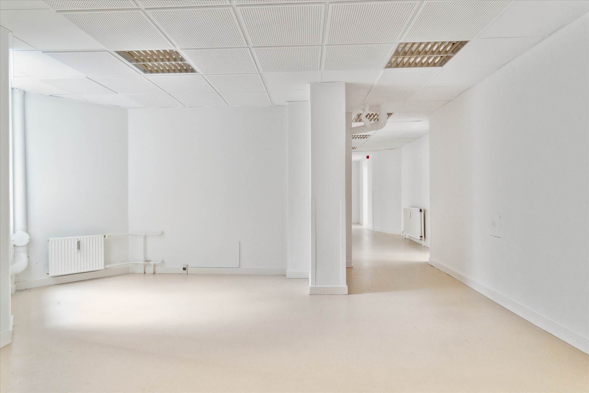Lys og rummelig kælderlejlighed med hvide vægge og gulv, højt loft med perforeret loftsbeklædning og radiatorer på væggen.