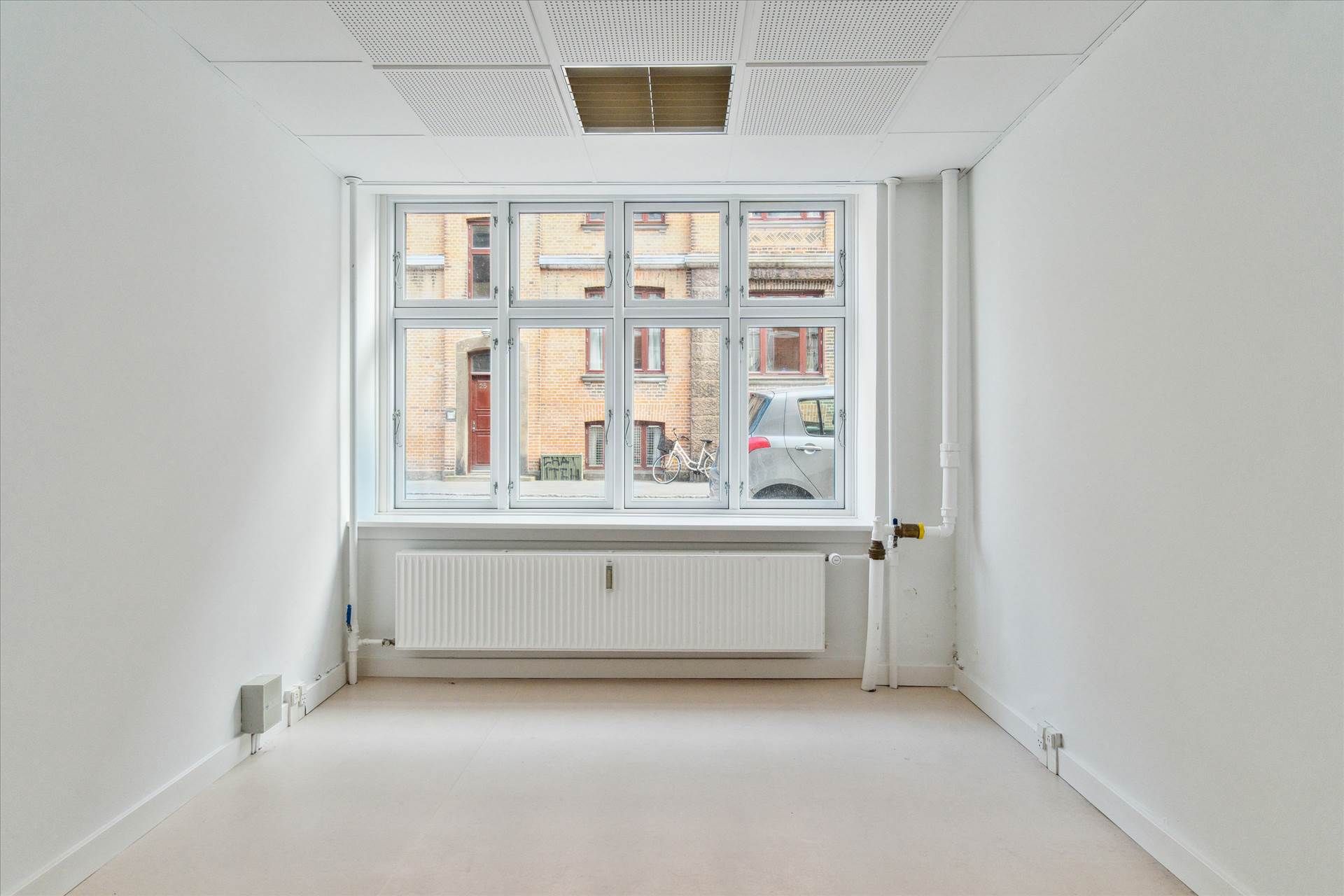 En hvid stue med to vindu og en radiator. På gaden udenfor er der en bil og en cykel.