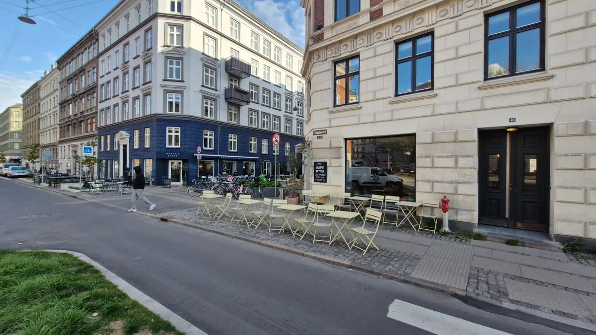 Café med grønne terrasseborde og stole udenfor Åboulevard 46, 2200 København N, med mand i sort tøj og hvid sko, der går på
