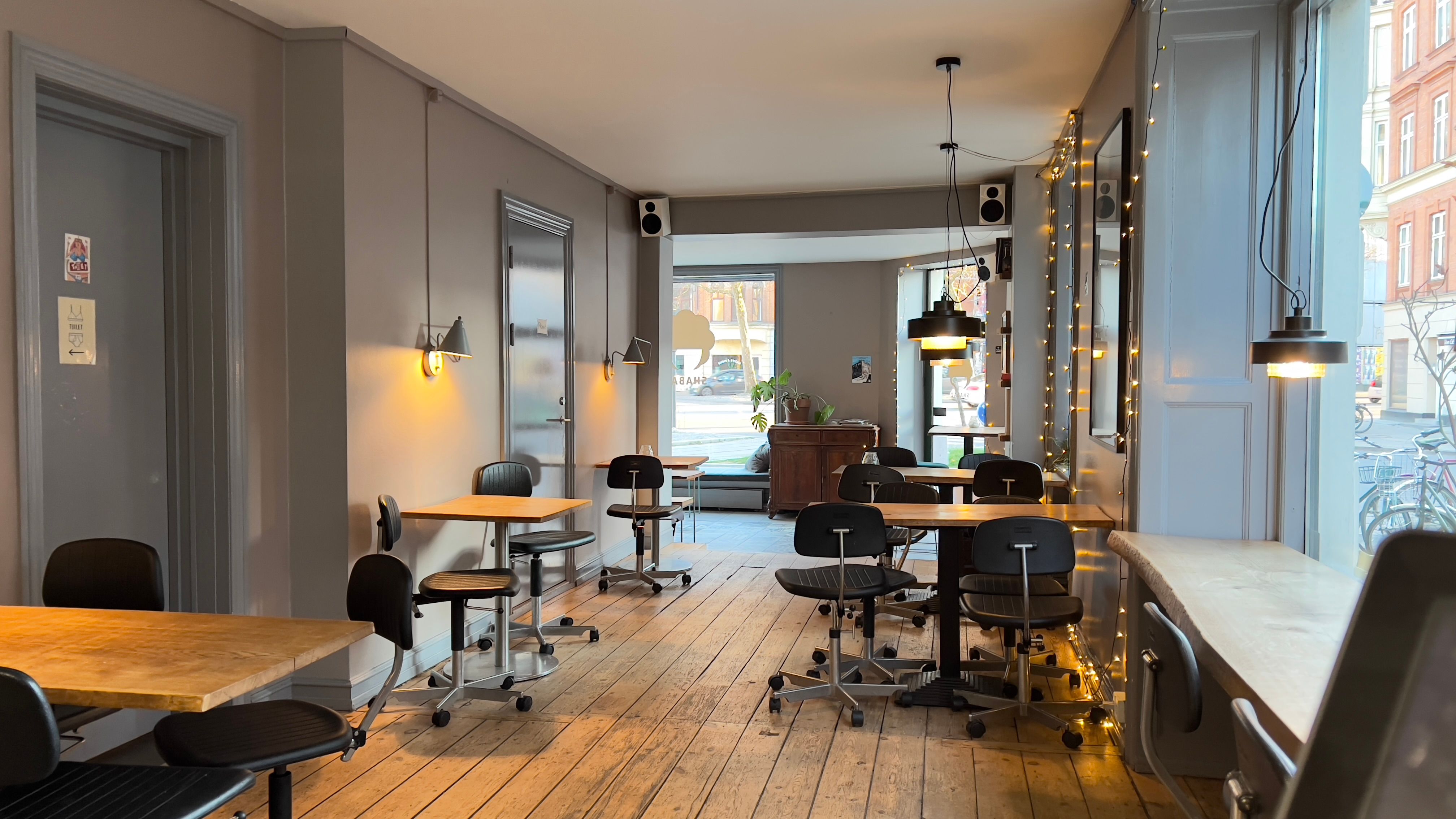 Møbler og lysekroner i en café med trægulv og hvide vægge samt udsigt til gågaden og cykler