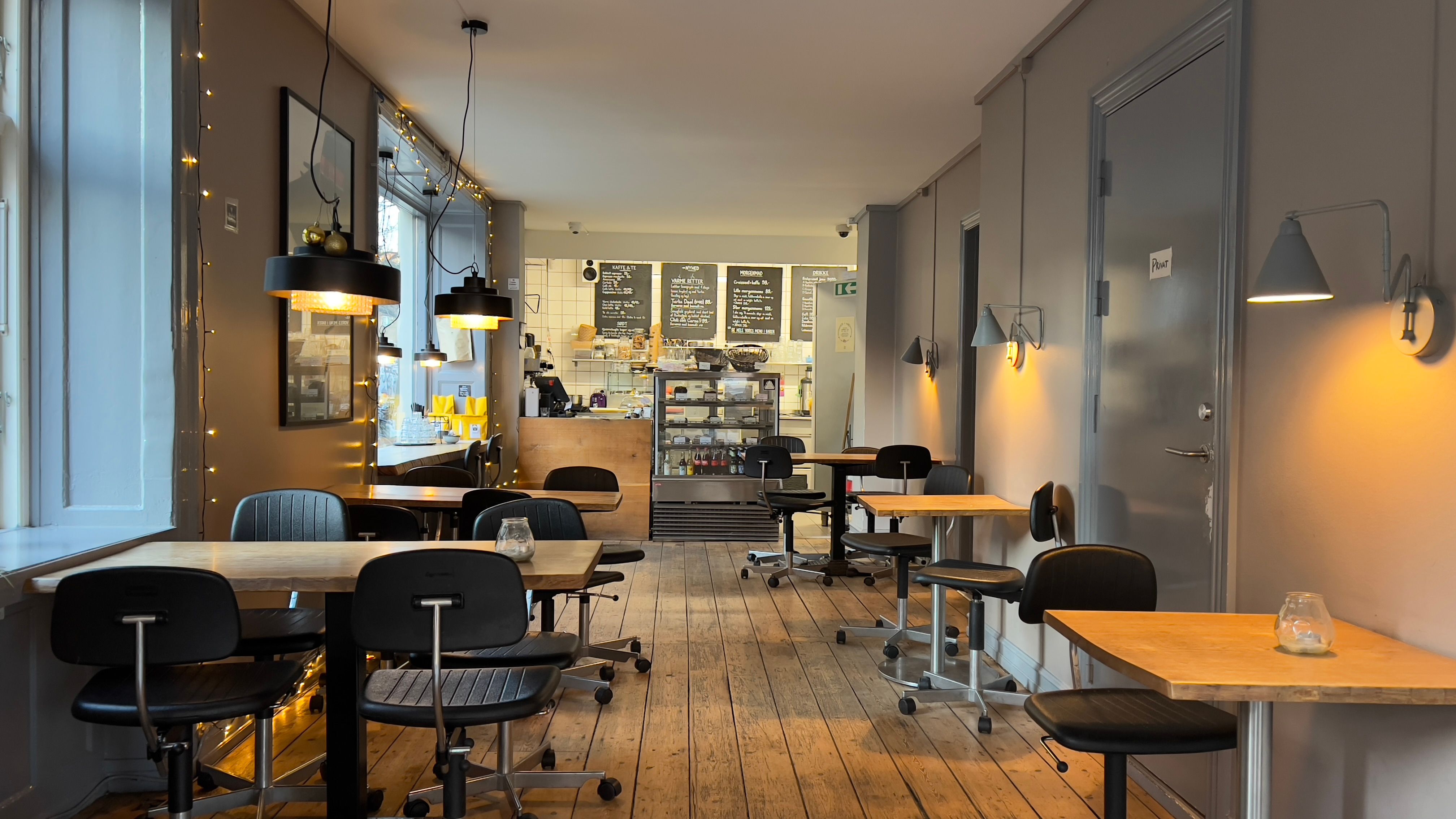 En moderne indrettet café med trægulve og lyse loftslamper, hvor borde og stole er anbragt i en åben planløsning