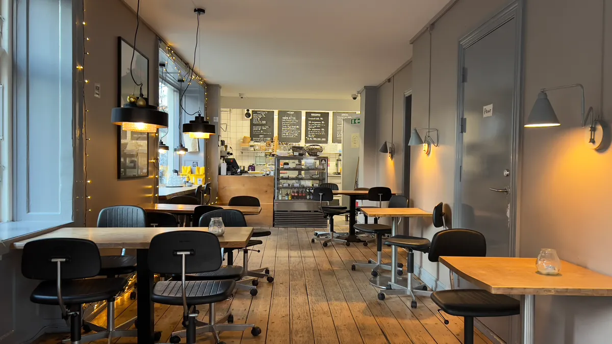 En moderne indrettet café med trægulve og lyse loftslamper, hvor borde og stole er anbragt i en åben planløsning