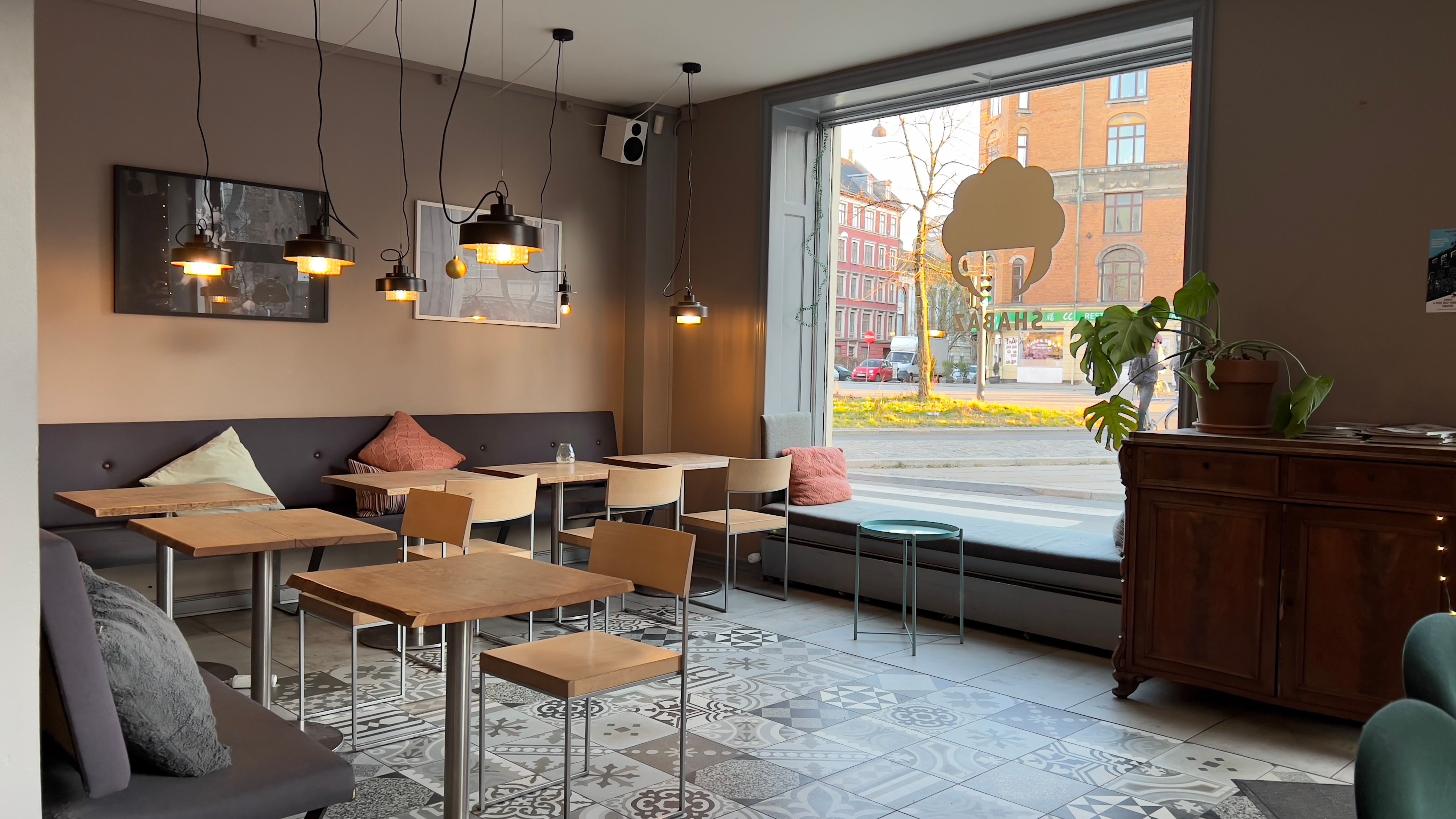 En café med bord og stole samt et vinduessæde med pude. Lyskroner hængende fra loftet og et skud af en bygade udenfor