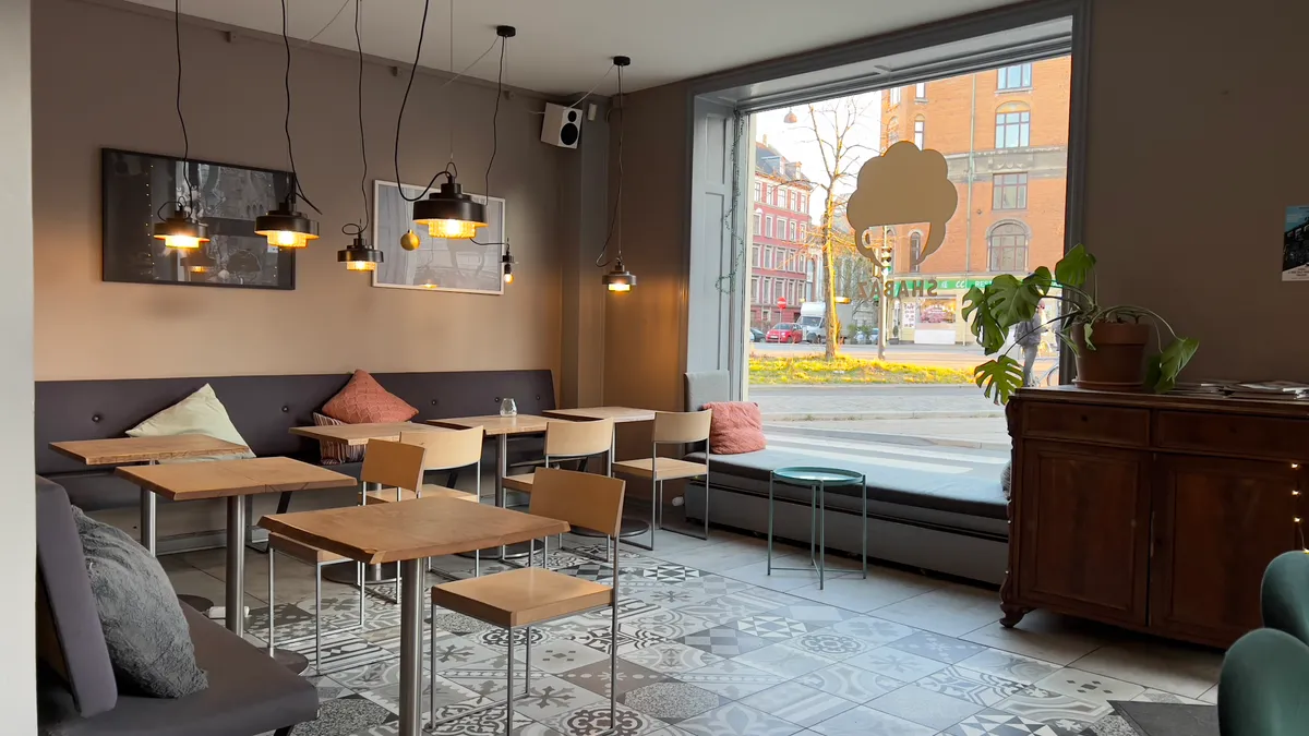 En café med bord og stole samt et vinduessæde med pude. Lyskroner hængende fra loftet og et skud af en bygade udenfor