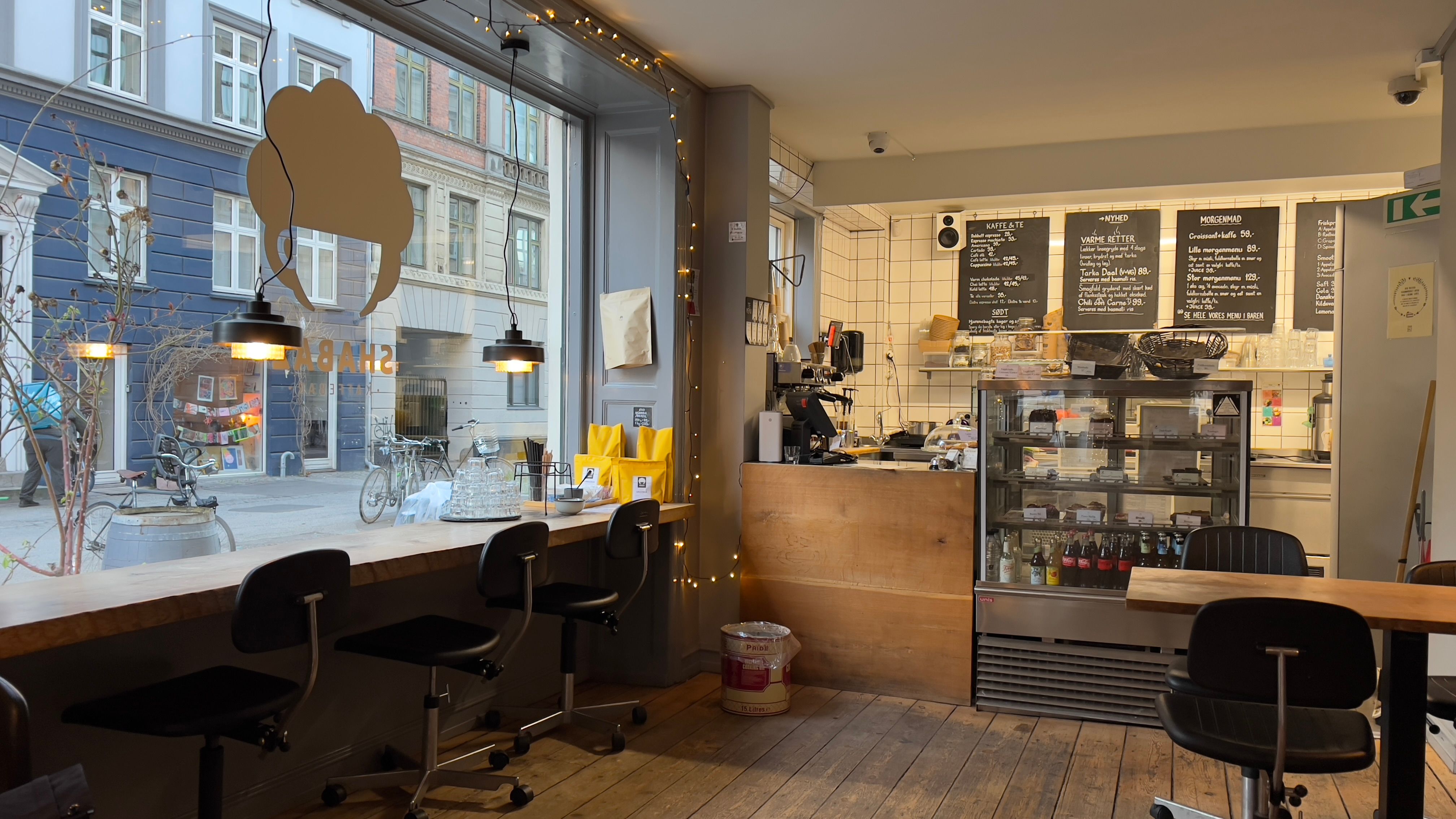 Store glasruder med lysekroner og hyggelige stole i en café med menuer og drikkevarer i skabet