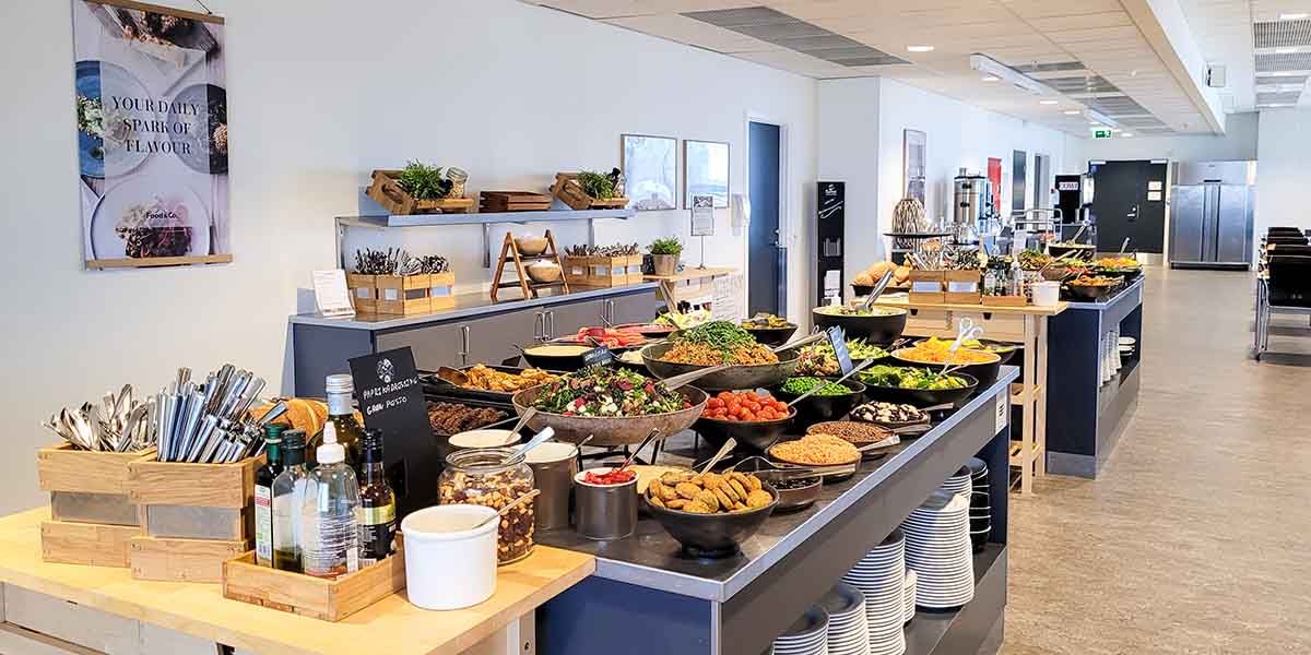 Buffet med salat, grøntsager, tomater, oliven, nødder, dressing og saucer samt drikkeflasker i et lyse kontorområde