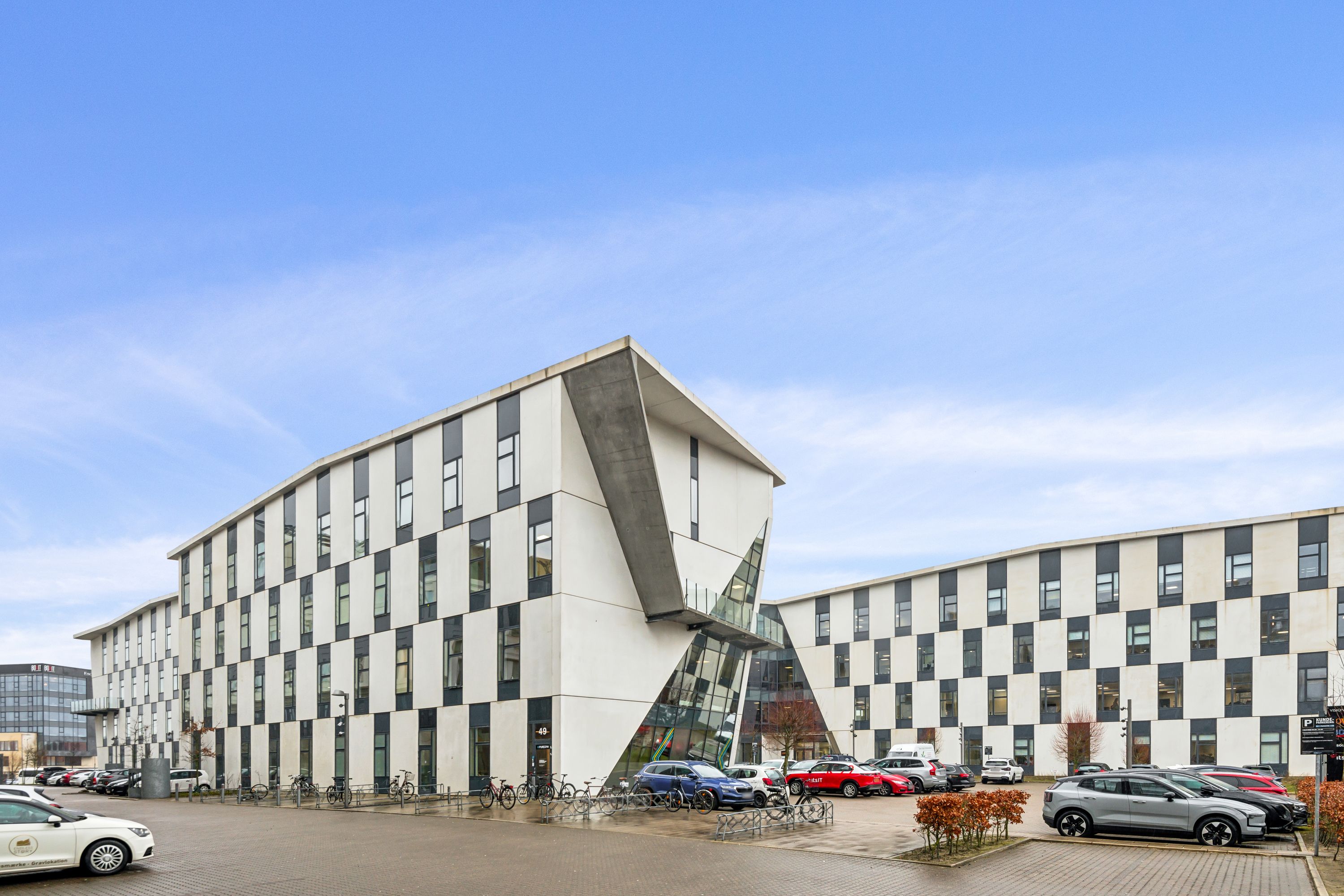 Moderne kontorbygning med glas og hvide og sorte vægge, flere parkerede biler, træer og planter, og et gult skilt med tekst