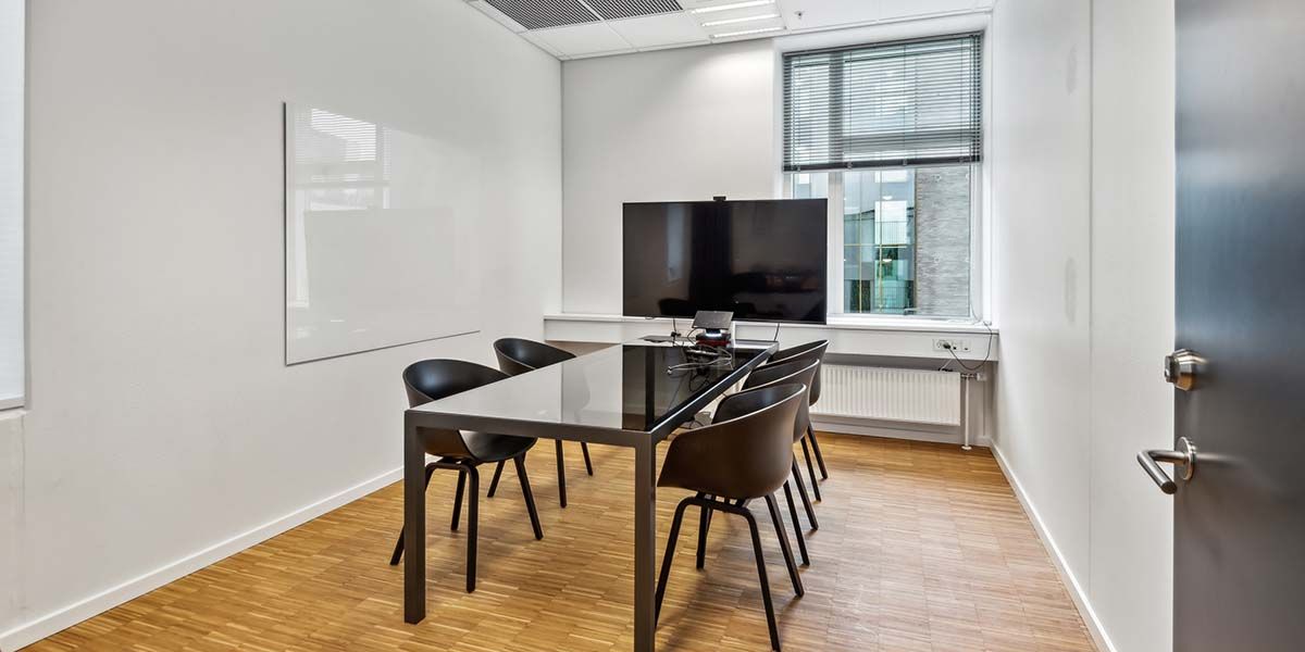 Moderne kontor med lyse vægge, trægulv og en stor tv-skærm foran et vindue med jalousier.