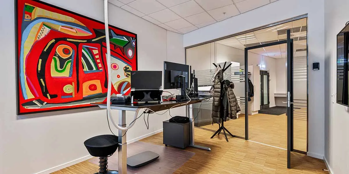 Kontorrum med moderne indretning, ståbord, skærm, kunstværk og tøj ved glasdør