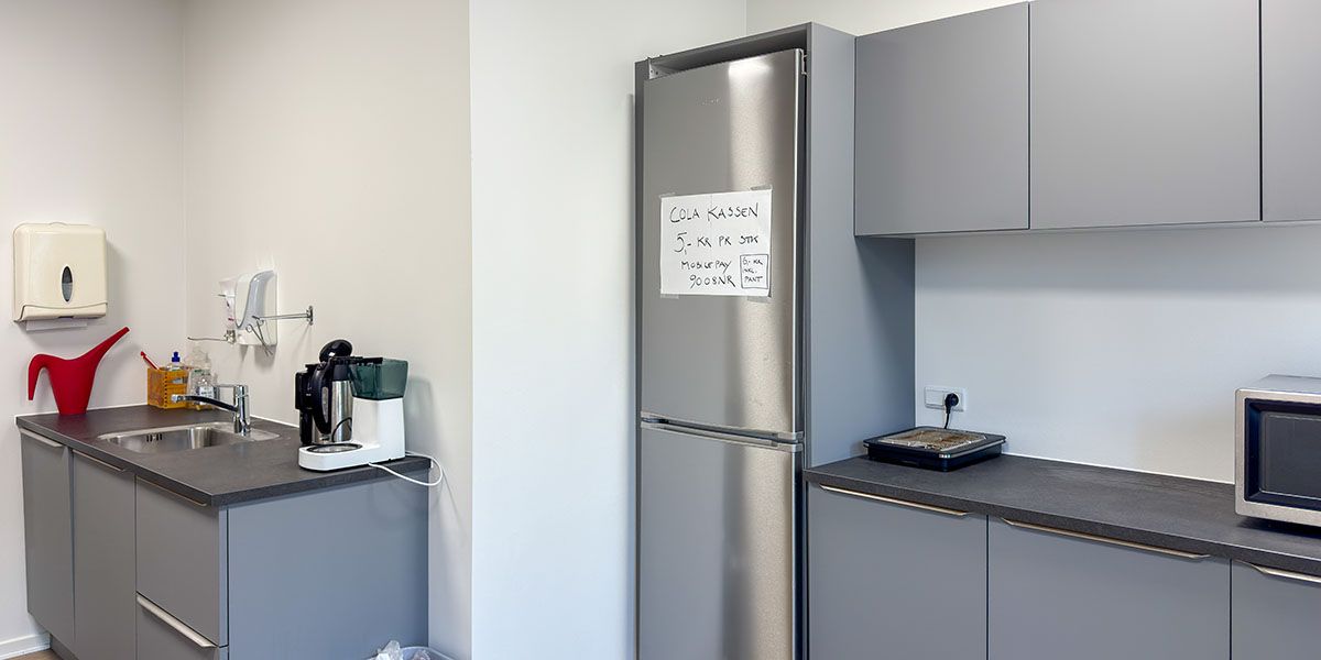 Funktionelt indrettet køkken med hvid væg og grå skabe, køleskab med skilt, mikroovn, kogeplade og kaffemaskine på bordpladen