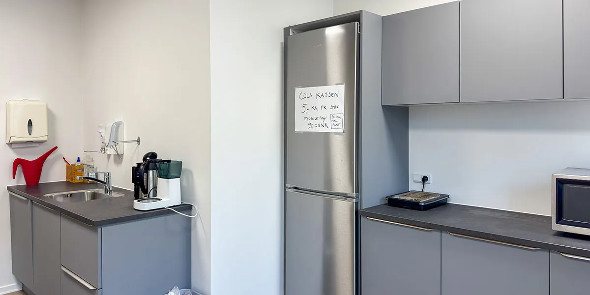 Funktionelt indrettet køkken med hvid væg og grå skabe, køleskab med skilt, mikroovn, kogeplade og kaffemaskine på bordpladen