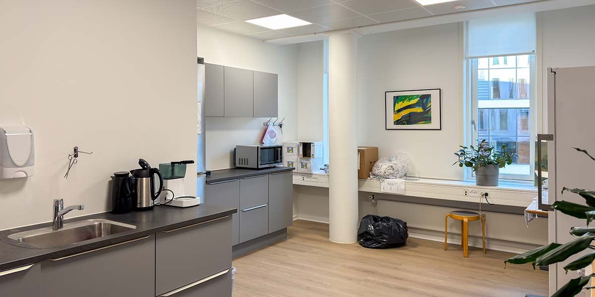 Køkken med en ovn, mikrobølgeovn, kaffemaskine, skraber og en håndsæbeautomat på væggen. Der er en stol, en kasse og en