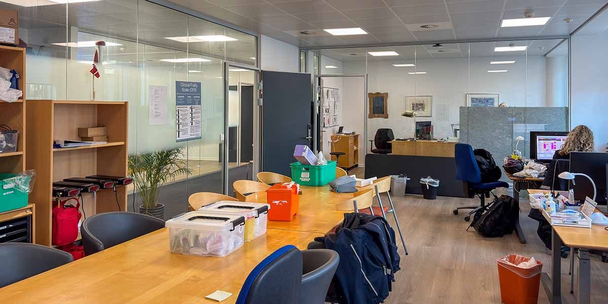 Kontor med mødelokaler og funktionelt indrettede kontorer med glasvægge og høje lofter. Bord med plastbokse og skove og en