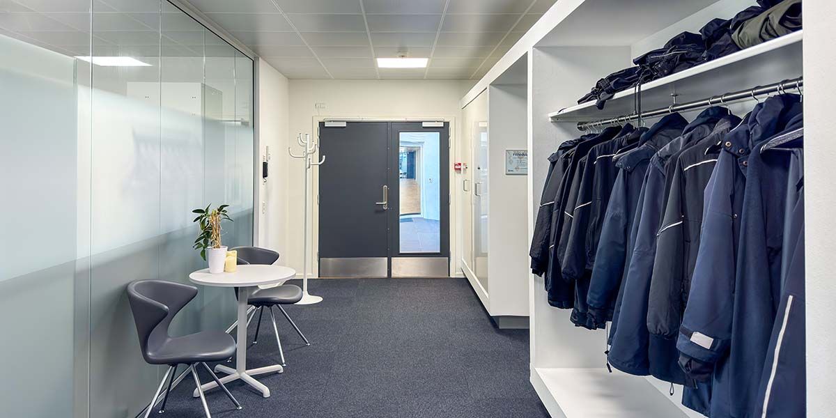 Moderne mødelokale med hvide vægge og glasvægge, møbler i lyse nuancer, en hvid rund bord og to stole, samt lyseblå frakker