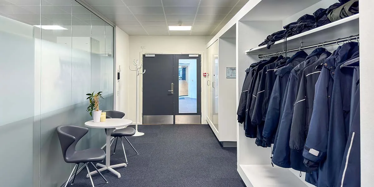 Moderne mødelokale med hvide vægge og glasvægge, møbler i lyse nuancer, en hvid rund bord og to stole, samt lyseblå frakker