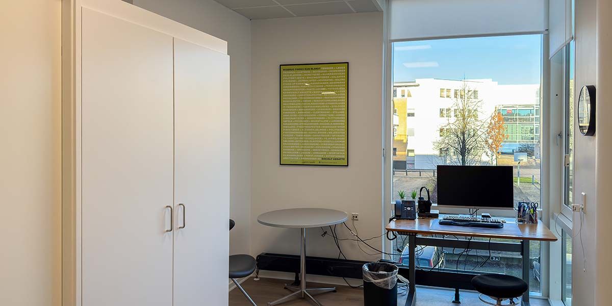 Kontor med computer, skrivebord, stol og højt vinduesparti. Et lysegult plakat hænger på væggen. Vinduet er åbent og ud til