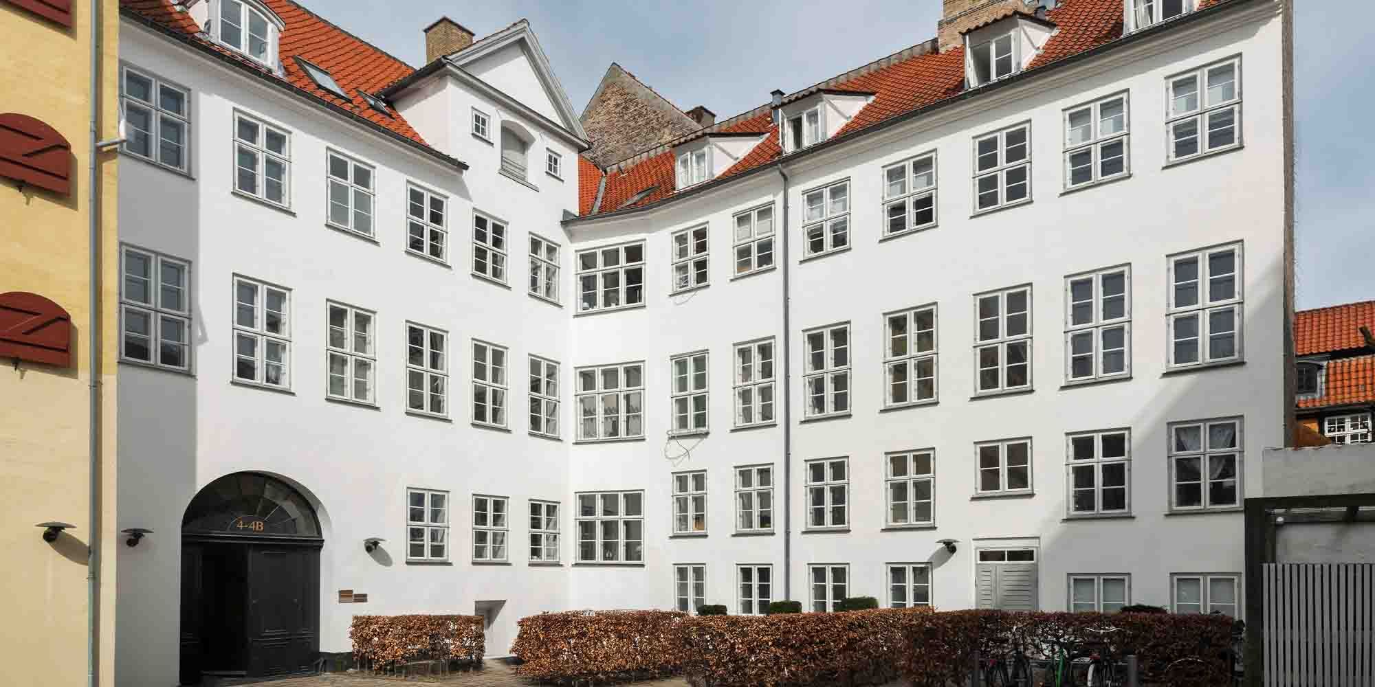 Bygning med lysegul facade, rødt tag og mange vinduer og indgange til lejemål på 1. sal med tre indgange