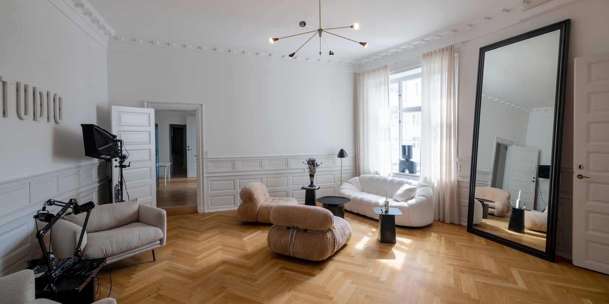 Et moderne interiør med lyse møbler og en stor spejl. Væggene er hvide og gulvet er træ. Et stort hvidt sovesofa med pude.