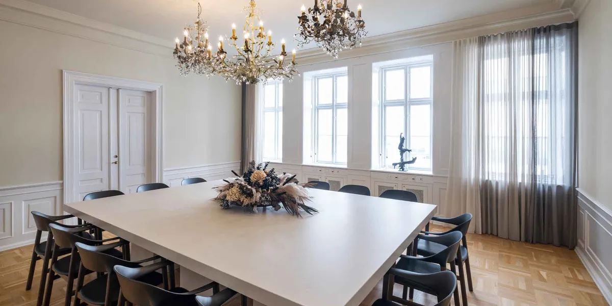 Langt bord med blomsterdekoration i mødelokale med hvide vægge og lysegult gulv samt to store lysekroner