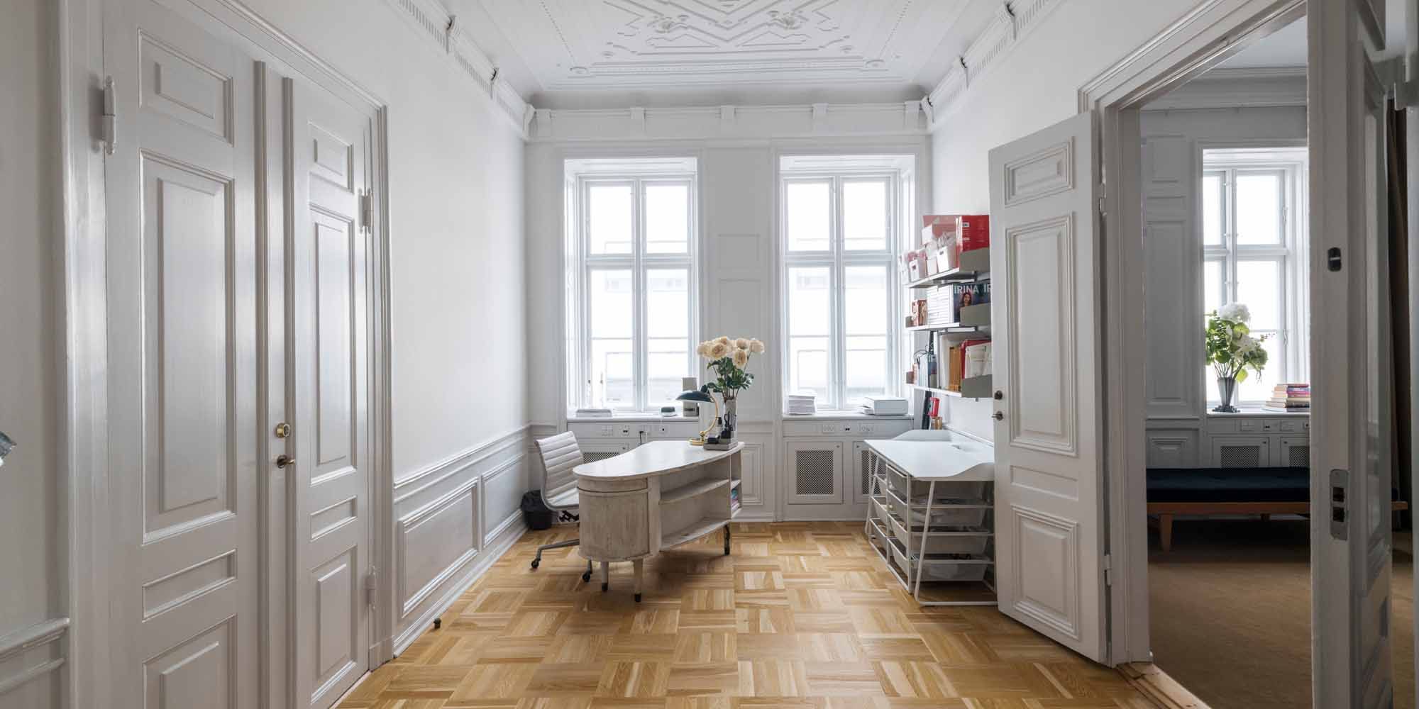 Et luksuriøst kontor med hvide vægge og lyse gulvfliser, to hvide skriveborde med lyse stole og et biblioteksreol med bøger