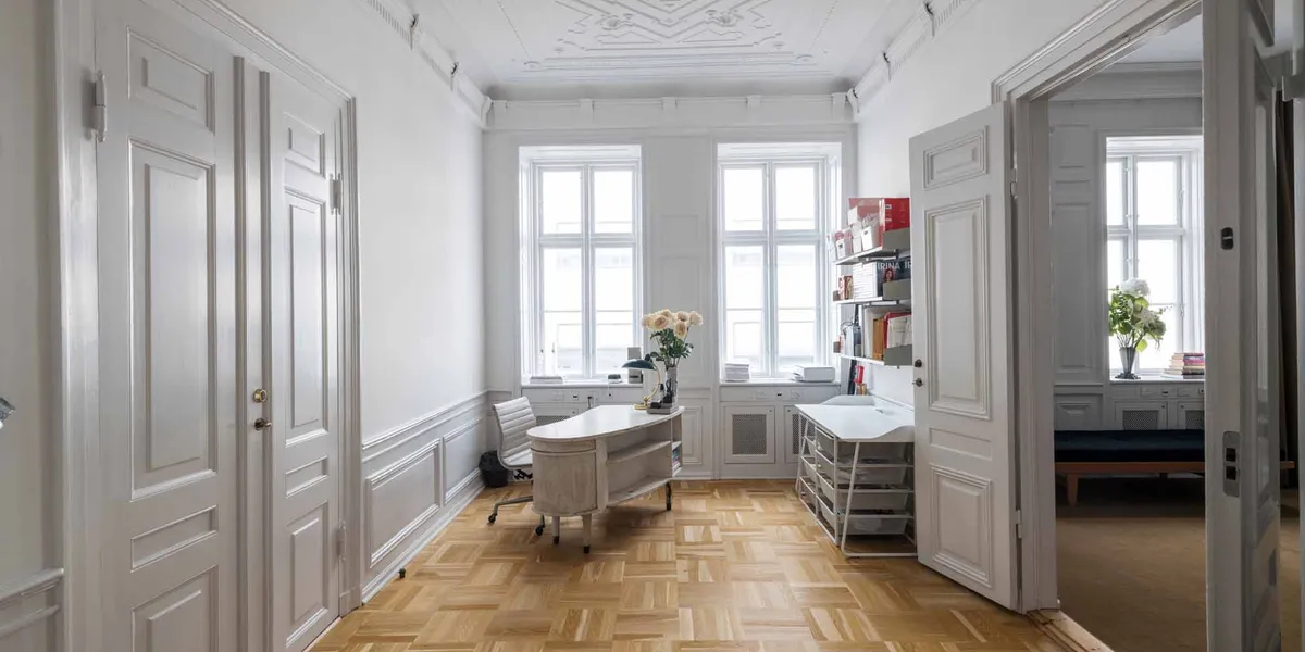 Et luksuriøst kontor med hvide vægge og lyse gulvfliser, to hvide skriveborde med lyse stole og et biblioteksreol med bøger