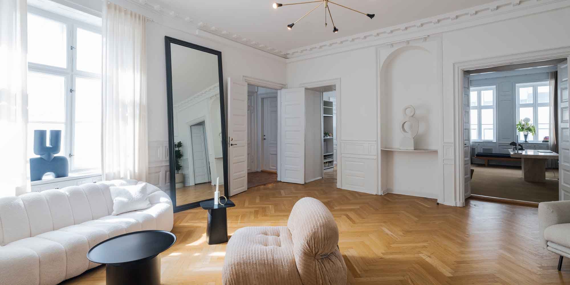 Lejlighed med højloft og møbler, som er i hvid og beige farve, samt en spejl og et bord med to lysstofrør