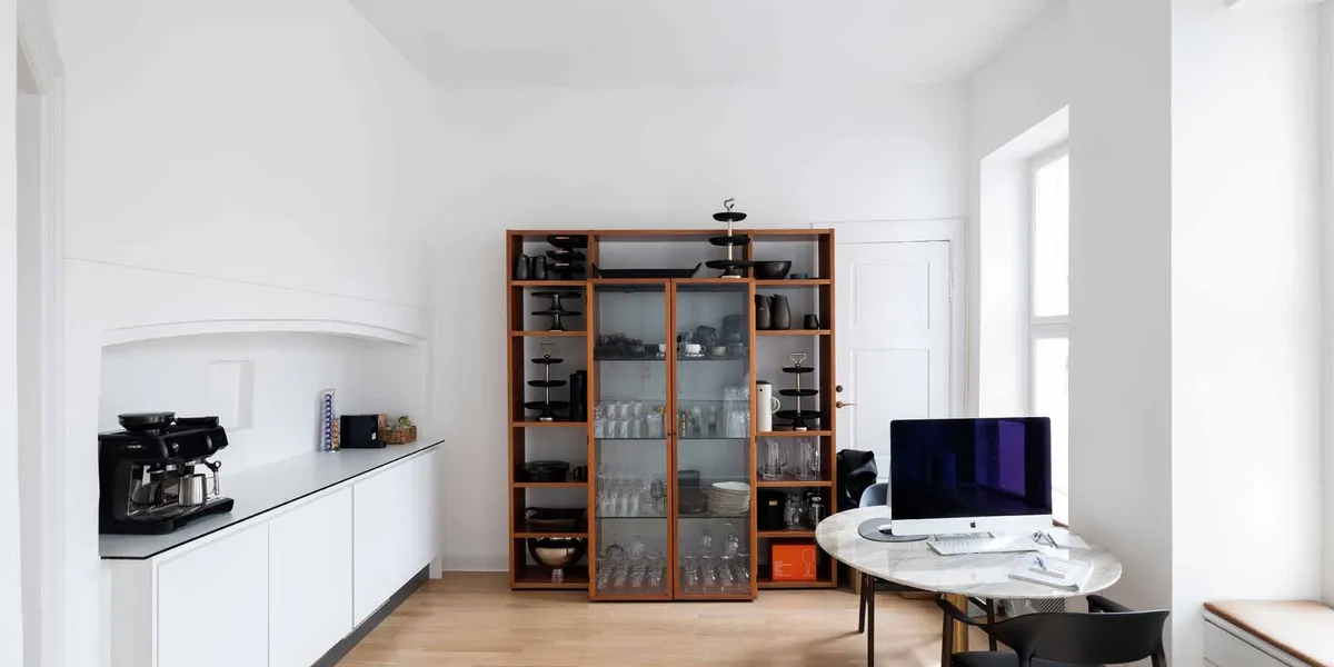 En lyse og minimalistisk køkken- og spisestue med en kaffemaskine, glaskabe og en rund marmorbord med computer.