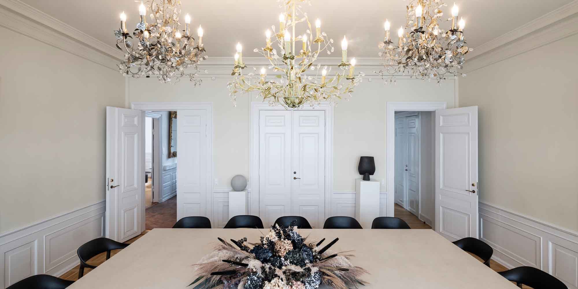 Store mødelokaler med lyse hvide vægge, store lysekroner, lyse møbler og en stor hvid bordplade med blomster.