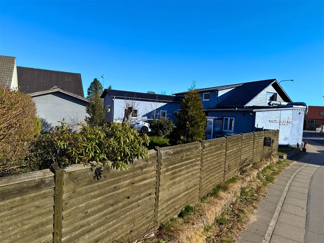 En hvid bil parkeret udenfor et blåt hus med røde tag, omgivet af en grøn plante og et højt gærde