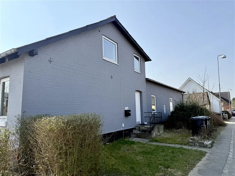 Et smukt hvidt hus med blå facade, fire vinduer og en skrånende tagkant, står i en grøn have og er fuld af græs og buske