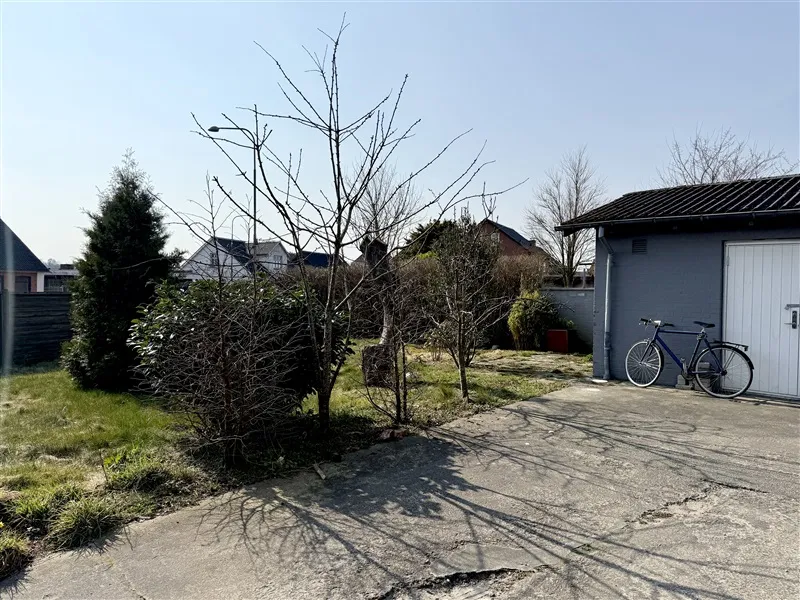 En cykel parkeret ved en garage, omgivet af buske, træer og græs, med baggrunden et hus og andre bygninger