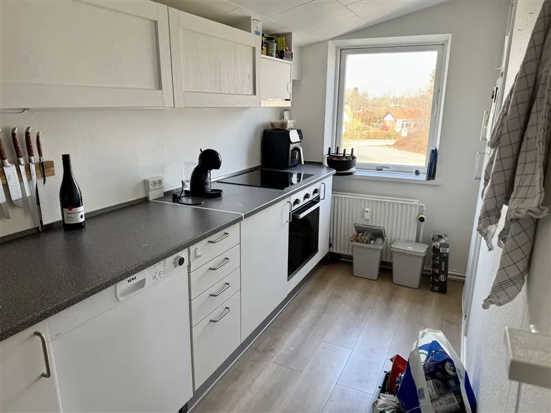 Et køkken med hvide skabe, sort bordplade, hvid radiator, to støvposer og en vinflaske