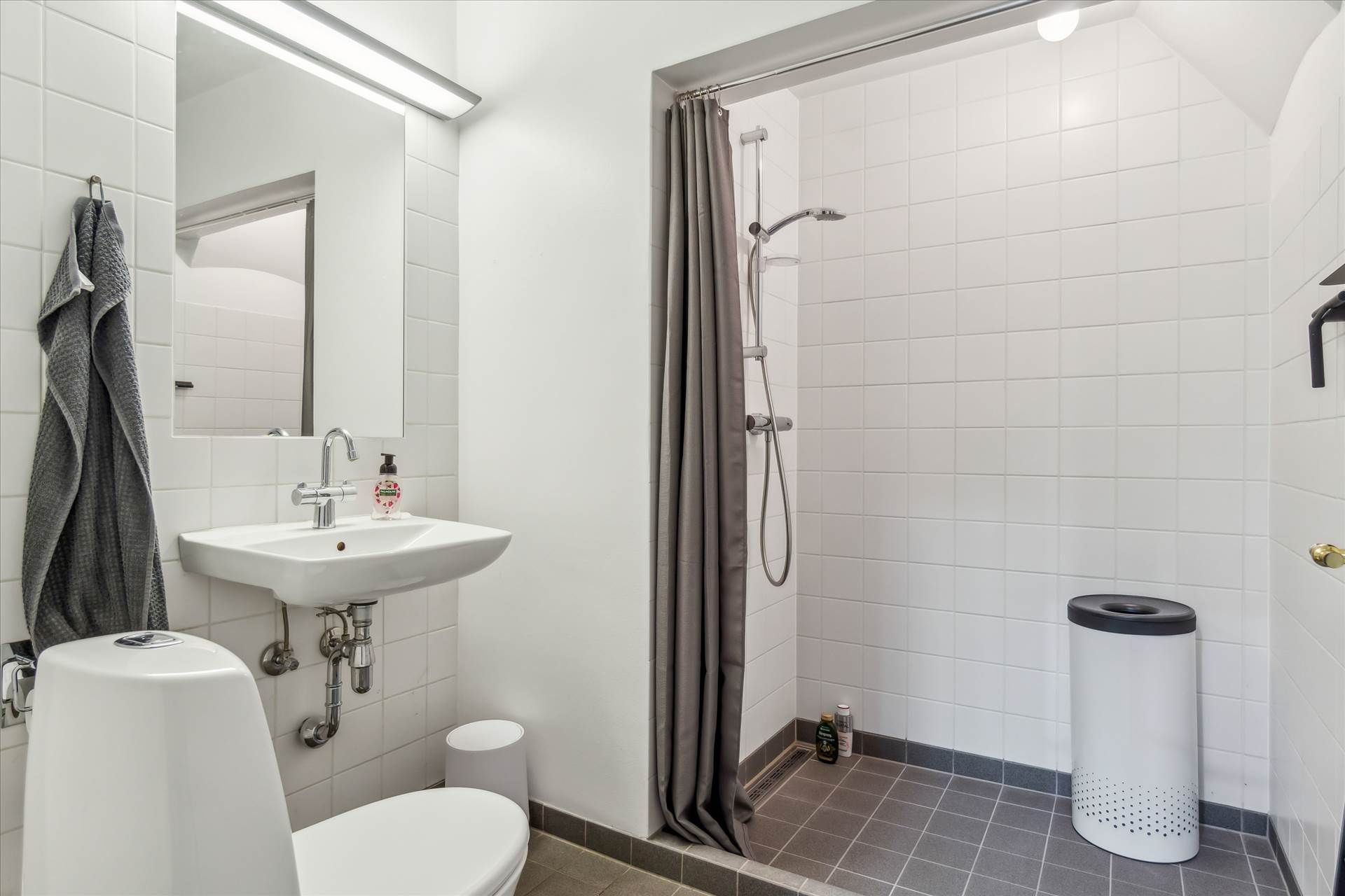 Et moderne hvidt badeværelse med brusebad, hvid toilet, hvidt spejl, hvidt håndvask, og en hvid papirkurv med sort låg